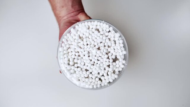 Adult hand holding a clear plastic container filled with small white polystyrene foam beads, top down view, clean white background