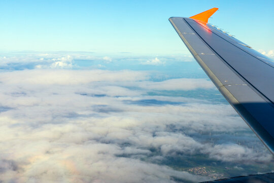 An aerial view from an airplane window, featuring the aircraft's wing and an orange winglet against a layered cloudscape and blue sky. Looking out over the leading edge of a commercial passenger jet.
