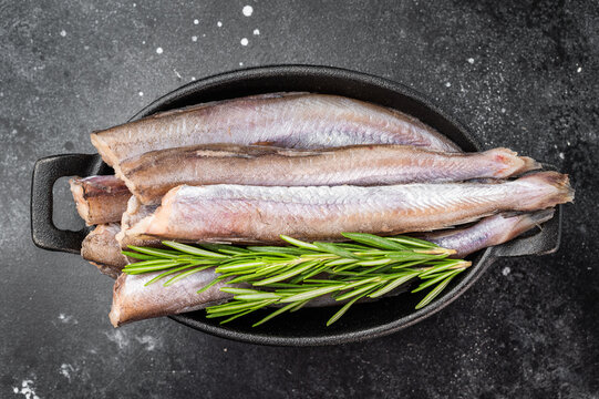 Uncooked blue whiting similar to small hake or codling arranged in black dish, natural marine seafood ingredient with herbs on dark stone kitchen background