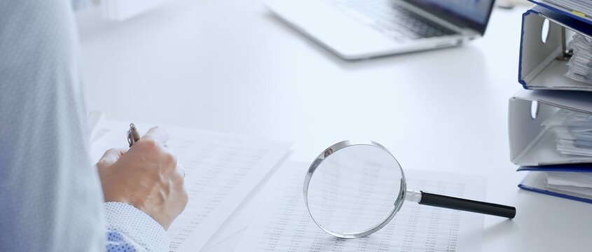 Accountants are analyzing financial reports with magnifying glass and documents, performing accounting and auditing tasks in the office, close up. Audit and taxes in business