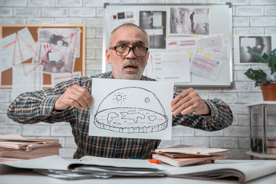 Senior flat Earth conspiracy theorist holding a hand drawn diagram of the world under a dome in front of an evidence board