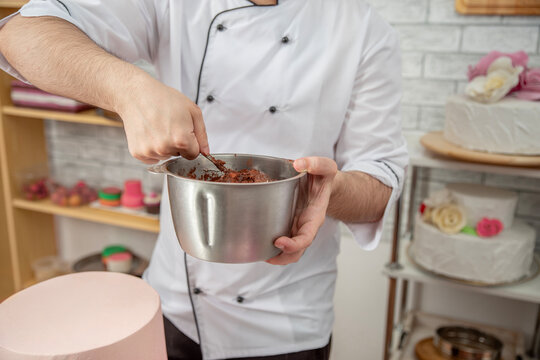 Close-up of hands of a pastry chef mixing chocolate cream with a spoon from a metal bowl in a confectionery workshop