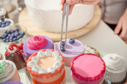 Hand of confectioner working on detailed dessert preparation at a worktable in a small artisan bakery kitchen. Pastry chef decorating miniature cakes with tweezers