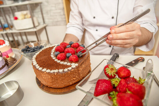 Pastry chef decorating a chocolate cake with fresh strawberries and blueberries using tongs in a confectionery workshop