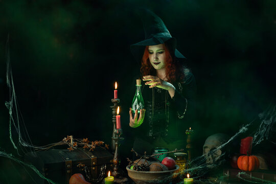 Witch conjuring a love potion, cooking up love, casts a spell, bottle with glowing poison in her hands. Young woman, a magician girl with red hair, dressed in Halloween attire wearing a conical hat