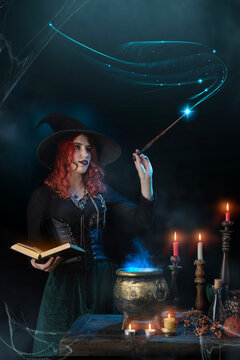 Witch with magic wand casts spell, cauldron, boiling magic potion, candles. Woman learns witchcraft from a book, magician girl with red hair, dressed in Halloween attire and conical hat