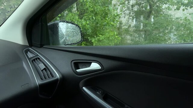 Interior of a car door with handle and vent, looking through a wet window with raindrops at the side mirror and green trees during a rainstorm.