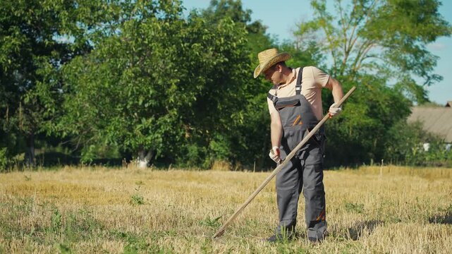 Male farmer in straw hat and gray overalls uses a hoe to cultivate dry grass in a sunlit field, surrounded by trees and a rustic building in the background