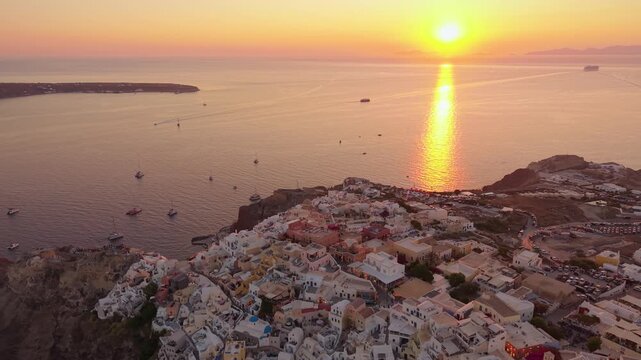 Panoramic drone of glowing sunset over Aegean Sea with Oia coastal town and boats in Santorini Greece