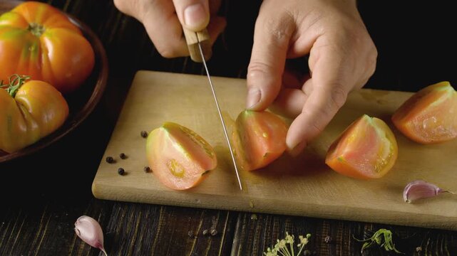 Hands slicing ripe tomatoes on a wooden cutting board with garlic cloves and herbs scattered around, showcasing the process of preparing fresh ingredients in a kitchen setting