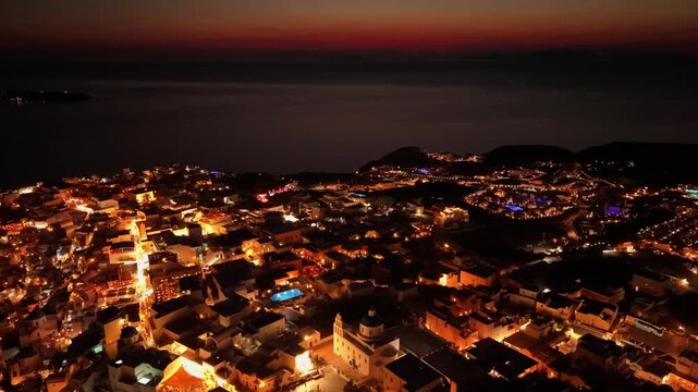 Night drone of Oia illuminated town lights and luxury resort pools under twilight sky in Santorini Greece