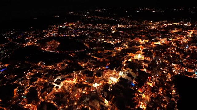 Panoramic night drone of sprawling Oia cliffside town illuminated under pitch black sky in Santorini Greece