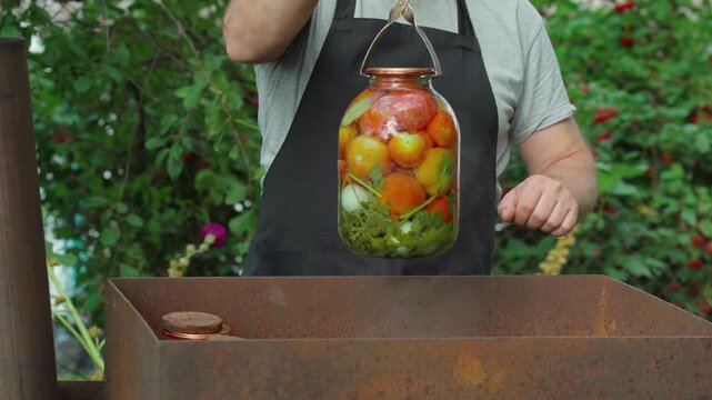 A cook in a black apron lifts a jar filled with bright tomatoes using metal tongs over a metal container. Canning vegetables outdoors
