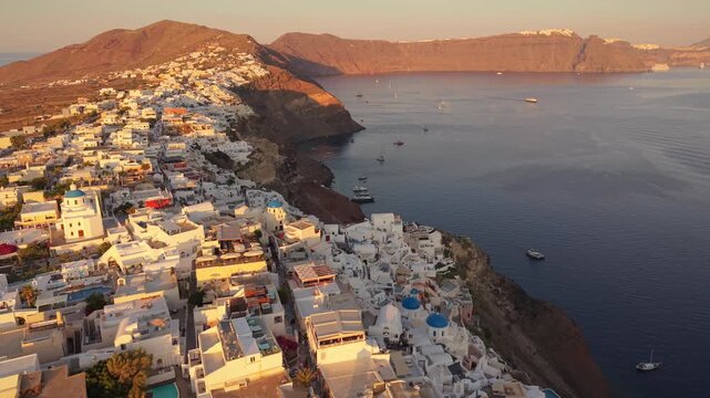 Aerial drone footage of Oia village perched on cliff edge at sunset with traditional white houses in Santorini Greece