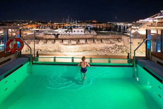 A cruise ship passenger enjoys views of the illuminated harbor and skyline of Rhodes, Greece, from a luxury ship deck's infinity pool at early evening.