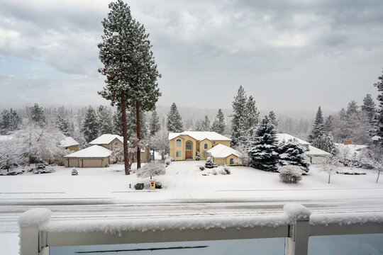 A scenic, snow-covered neighborhood view overlooking a residential street of homes surrounded by tall pine trees under a heavy overcast sky in Coeur d'Alene, Idaho.