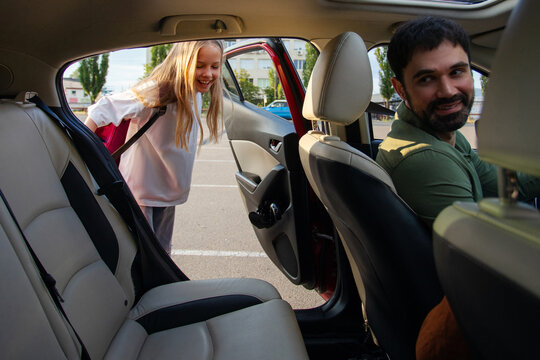 Schoolgirl with backpack getting into car while her father turning to her and smiling during warm afternoon school pickup, view from inside