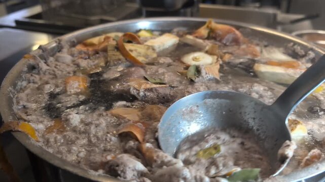 Close-up of a chef skimming grey foam and impurities off the surface of a simmering beef broth using a ladle. The pot contains beef, root vegetables, onion skins and bay leaves, showing the traditiona