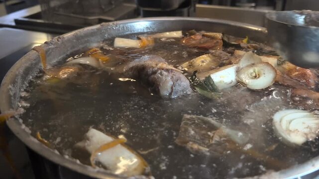 Close-up of a chef skimming grey foam and impurities off the surface of a simmering beef broth using a ladle. The pot contains beef, root vegetables, onion skins and bay leaves, showing the traditiona