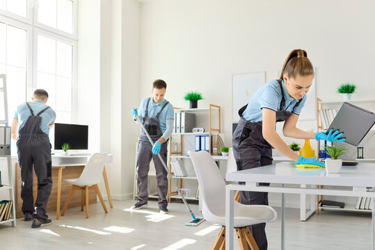 Cleaning team disinfecting office workspace with supplies. Uniformed cleaners sanitize desks with gloves and spray for hygiene. Concepts include cleanliness and professional service.