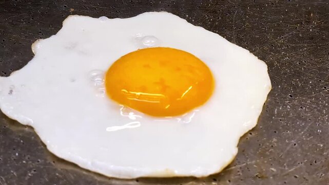 Close-up shot of a sunny side up fried egg with a bright orange yolk cooking on a hot flat top griddle. The egg white sizzles and sets while the golden yolk remains glossy and intact, capturing the cl