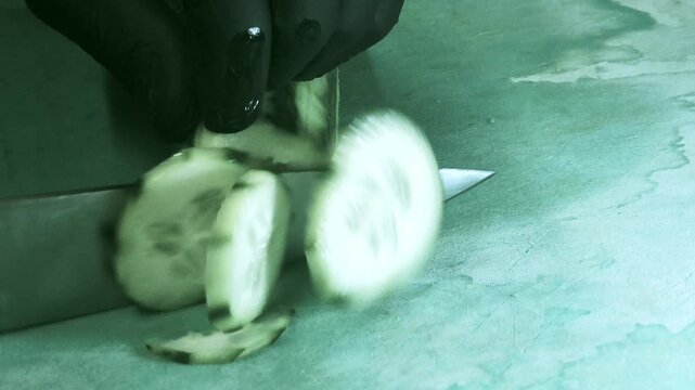 Close-up of a chef's gloved hand slicing a fresh cucumber into thin round slices with a sharp knife on a green cutting board. The slices fall away one by one, showing crisp texture and seeds, ideal fo