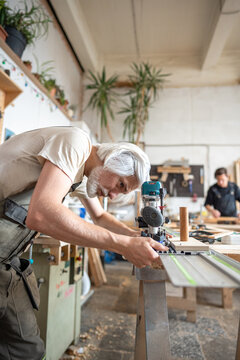 Attentive carpenter carefully adjusts wood router machine with guide rail adapter at workbench in studio, craftsman checks accuracy of router with metal track to repair vintage wooden door in workshop