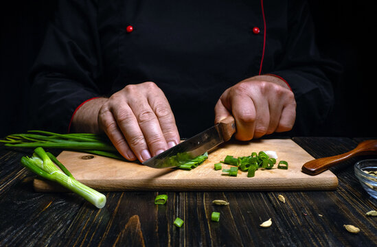 A chef is chopping green onions on a wooden cutting board in a kitchen. The chef focuses on the task while sliced green onions are placed around