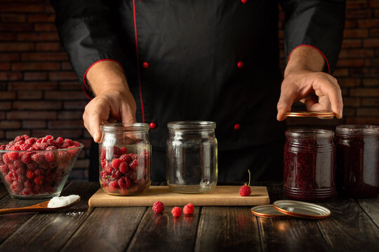 Chef is working in a kitchen, placing fresh raspberries into a glass jar. On the table, there are more raspberries in a bowl and jars containing jam