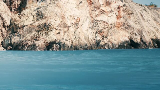 A rocky cliff stands tall beside a large body of water. The sun shines on the surface of the water, creating ripples. Trees are scattered on the cliff in the background.