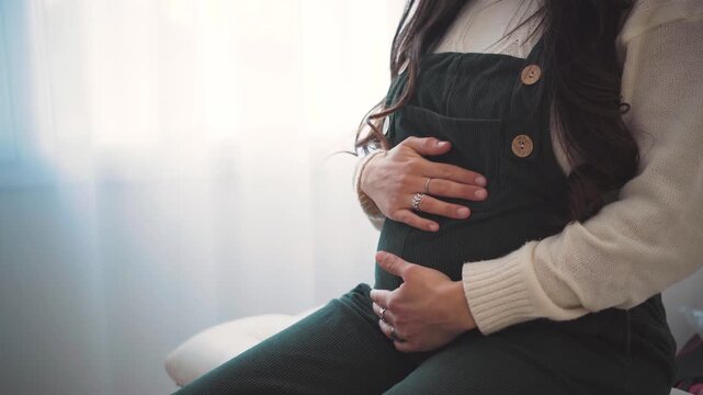 Close-up of an anonymous pregnant young woman's hands gently resting on and caressing her belly.

