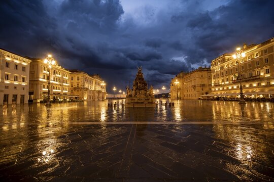 Illuminated square Piazza Unit&agrave; d'Italia with Fontana dei Quattro Continenti fountain, Palazzo del Lloyd Triestino and Palazzo della Luogotenenza Austriaca or Palazzo Governo, with reflection, dramatic clouds, blue hour, old town, Trieste, Friuli-Venezia Giulia region, Italy