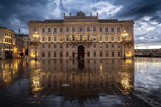 Illuminated square Piazza Unit&agrave; d'Italia, Palazzo del Lloyd Triestino, with reflection, dramatic clouds, blue hour, old town, Trieste, Friuli-Venezia Giulia region, Italy