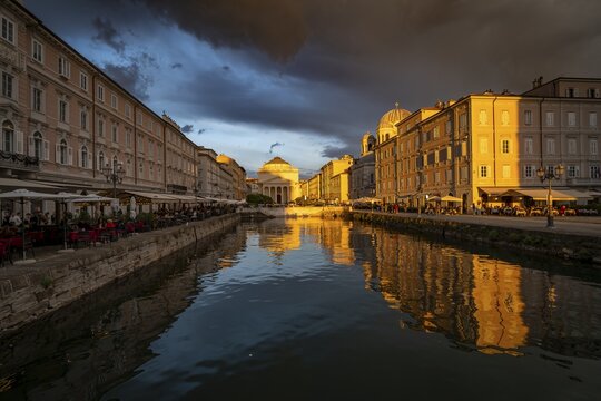 Grand Canal with restaurants, Sant'Antonio Taumaturgo church and Serbian Orthodox church Santissima Trinit&agrave; e di San Spiridione, reflection in the canal, in the evening light at sunset, dramatic clouds and atmospheric lighting, Borgo Teresiano, Trieste, Friuli-Venezia Giulia region, Italy