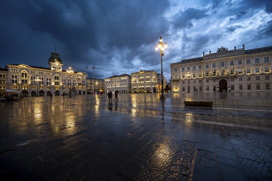Illuminated square Piazza Unit&agrave; d'Italia, City Hall, Palazzo Communale and Palazzo del Lloyd Triestino, with reflection, dramatic clouds, blue hour, old town, Trieste, Friuli-Venezia Giulia region, Italy