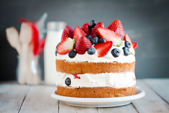 Close-up of a Sponge layer cake with fresh strawberries, blueberries and cream on a wooden table with a bottle of milk and cutlery