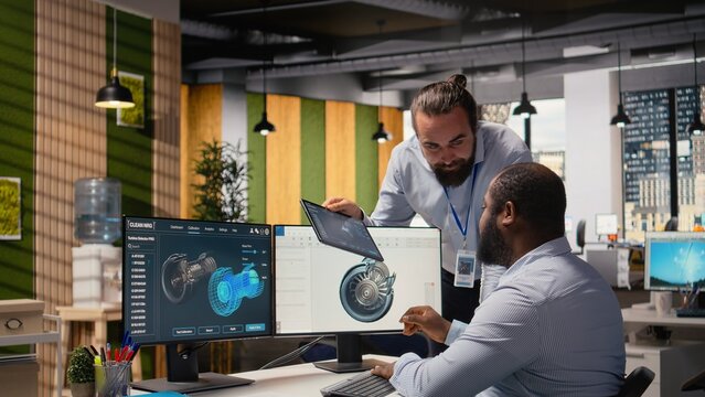 Team of technicians works together to upgrade turbine parts for sustainability while colleagues use CAD design software to brainstorm blade aerodynamics and boost green energy output in the office.