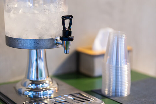 Close-up view of a beverage dispenser filled with cold iced water or juice, featuring a metallic chrome stand and spigot Stacked clear plastic cup are placed in background, event, or cafe setup.