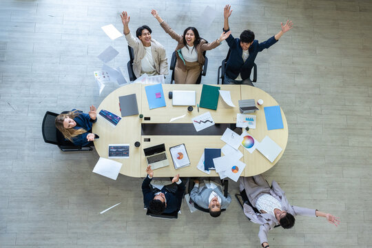 Top view of excited business team around a large conference table, cheering and throwing papers into the air to celebrate successful project completion, freedom, or achievement in a modern office.