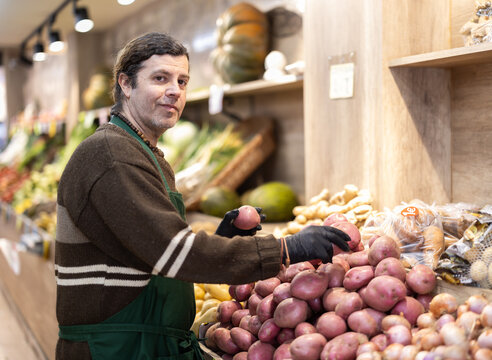Adult male seller in uniform lays out potatoes on counter in vegetable store