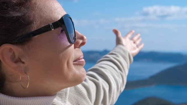 Woman with outstretched arms enjoying the sunny day and sea view. Happy woman wearing sunglasses looking up with a smile and enjoying the sunshine, her arm outstretched towards the sea landscape