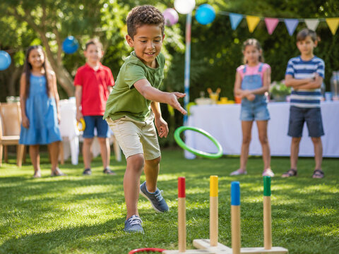 Energetic boy throws a green ring toward a colorful ring-toss game at a sunny backyard party while friends watch from the softly blurred background.