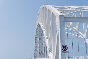 White steel bridge curves gracefully against clear blue sky. Strong structural beams form elegant, geometric pattern. Modern engineering meets natural sky in striking contrast. Ideal for architecture © Rizky Rahmat Hidayat