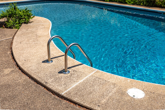 Outdoor swimming pool with metal ladder, blue water and curved concrete deck in summer sunlight