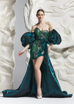 Full length shot of elegant woman wearing luxury emerald green balloon sleeve cocktail mini dress with sweeping draped train posing gracefully against sculptural white studio wall backdrop