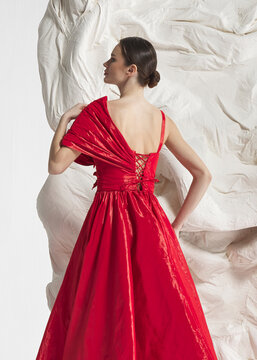 Rear back view of elegant woman wearing luxury crimson red metallic red taffeta ballgown with sculptural rosette and open back posing gracefully against sculptural white studio wall backdrop