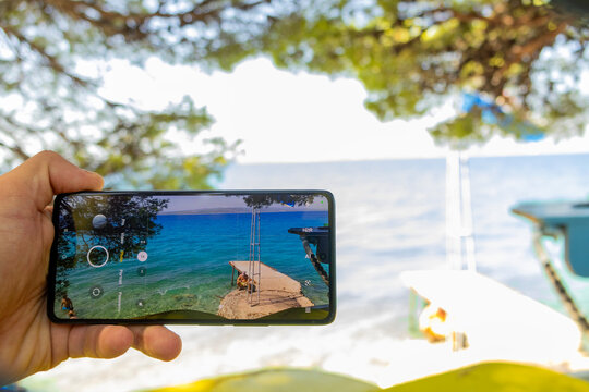 Hand holding a smartphone while photographing a seaside pier extending into turquoise water, with a bright coastal landscape creating a summer travel and vacation scene.