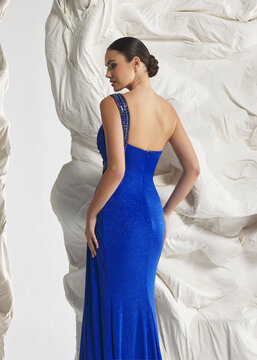 Rear back view of elegant woman wearing luxury royal blue glitter draped jersey evening gown with open back and cowl drape posing gracefully against sculptural white studio wall backdrop
