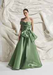 Fototapeta premium Full length shot of elegant woman wearing luxury sage green strapless satin ballgown with oversized bow at waist and wide hem posing gracefully against sculptural white studio wall backdrop