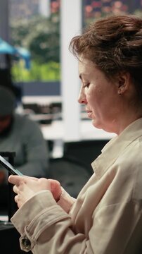 Portrait format: Smiling woman texts on a touchscreen smartphone in a bus station while waiting for an intercity coach connection, chatting online with friends on her mobile phone before departure.